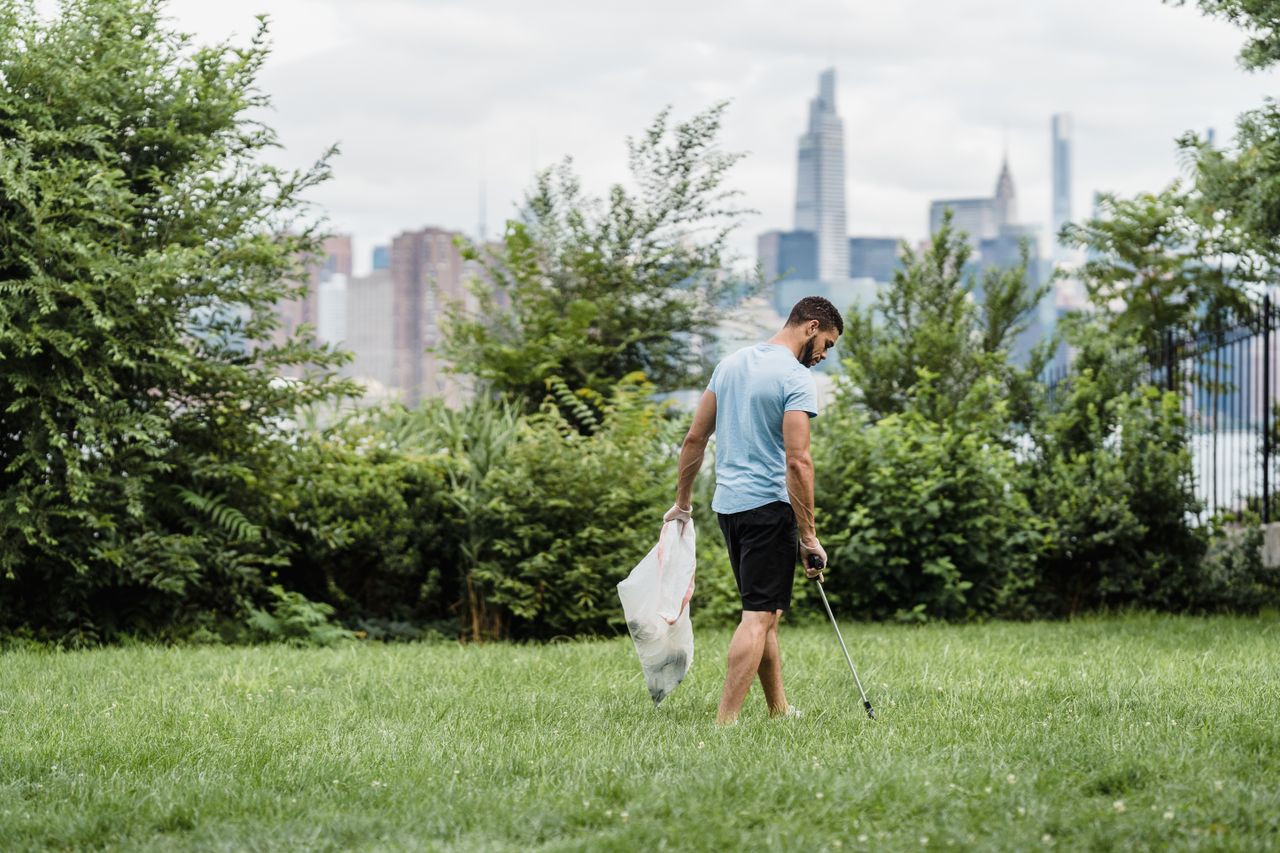 hero-img-01 Man picking up litter in a park with the city skyline in the background, promoting environmental conservation.